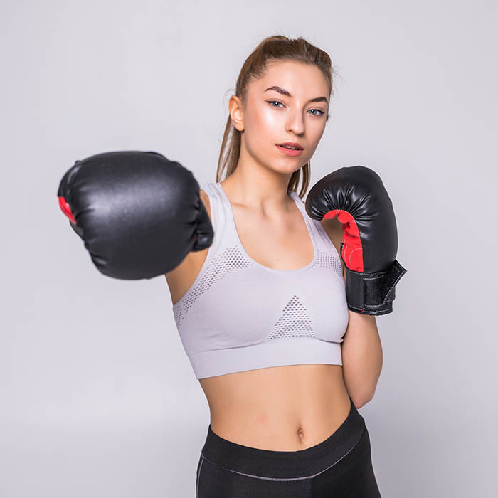 Portrait of young woman boxer throwing a punch at camera while practicing on grey background. Mixed race female athlete wearing boxing gloves exercising boxing. Portrait of a young woman boxer throwing a punch at camera while practicing on grey background. Mixed race female athlete wearing boxing gloves exercising boxing.